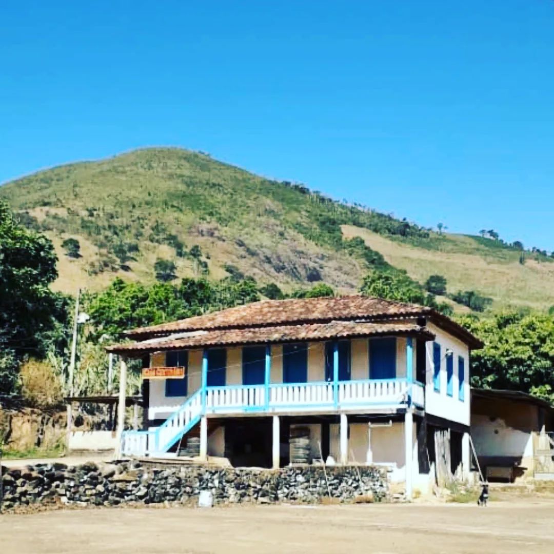 A rustic house is positioned at the base of a green hillside, featuring a traditional tiled roof and blue accents on the windows and balcony. The landscape is open, with a clear blue sky above, creating a serene backdrop.
