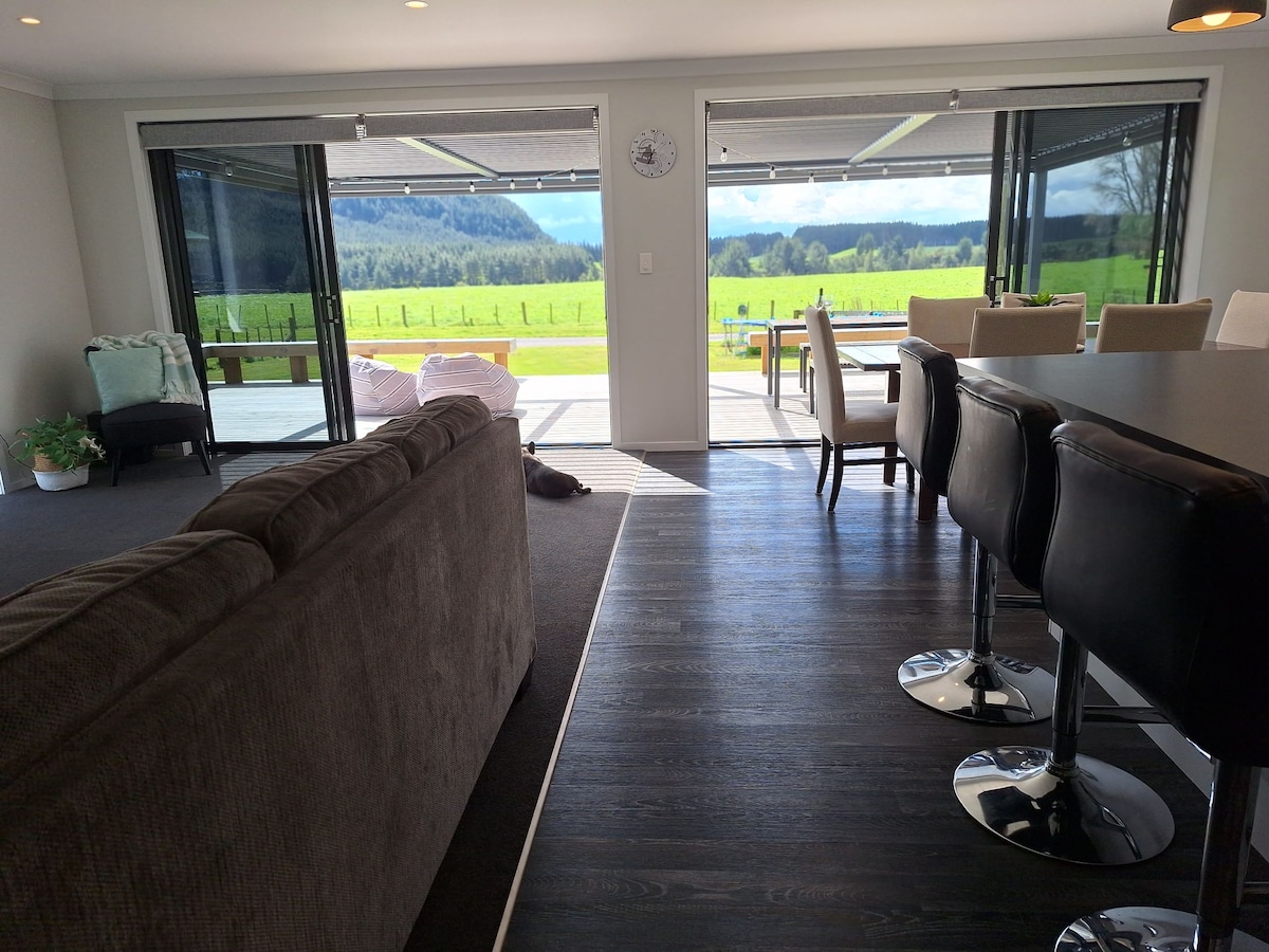 A spacious open-plan area is showcased, featuring a comfortable couch and a dining table. Glass sliding doors provide a clear view of the vibrant green rural landscape outside, seamlessly connecting indoor and outdoor spaces. Natural light fills the area, enhancing the welcoming environment.