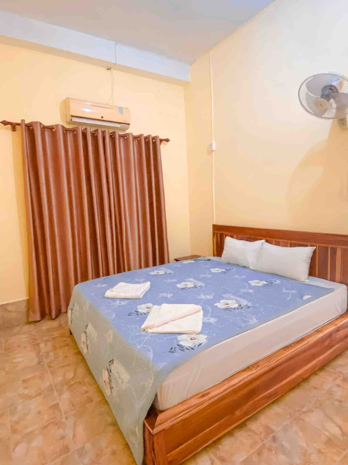 A cozy sleeping area features a wooden bed with a blue floral bedspread, complemented by crisp white pillows. Soft light flows through the partially closed curtains, while an air conditioning unit is mounted on the wall, ensuring comfort in the space.