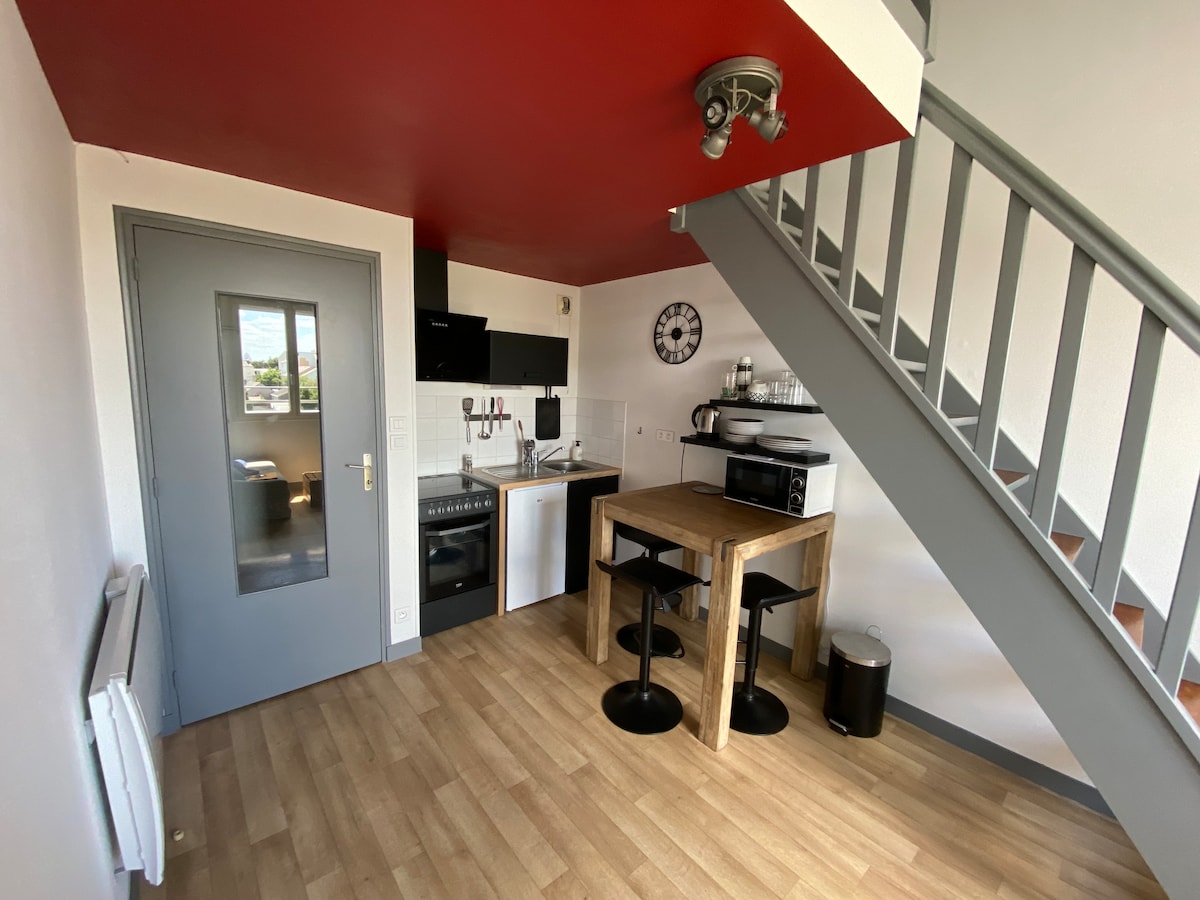 The compact kitchen area features modern appliances, including a refrigerator, stove, and microwave. A wooden table with two stools is positioned near a window, while a staircase leads to the upper level. A clock adds a practical touch to the wall.