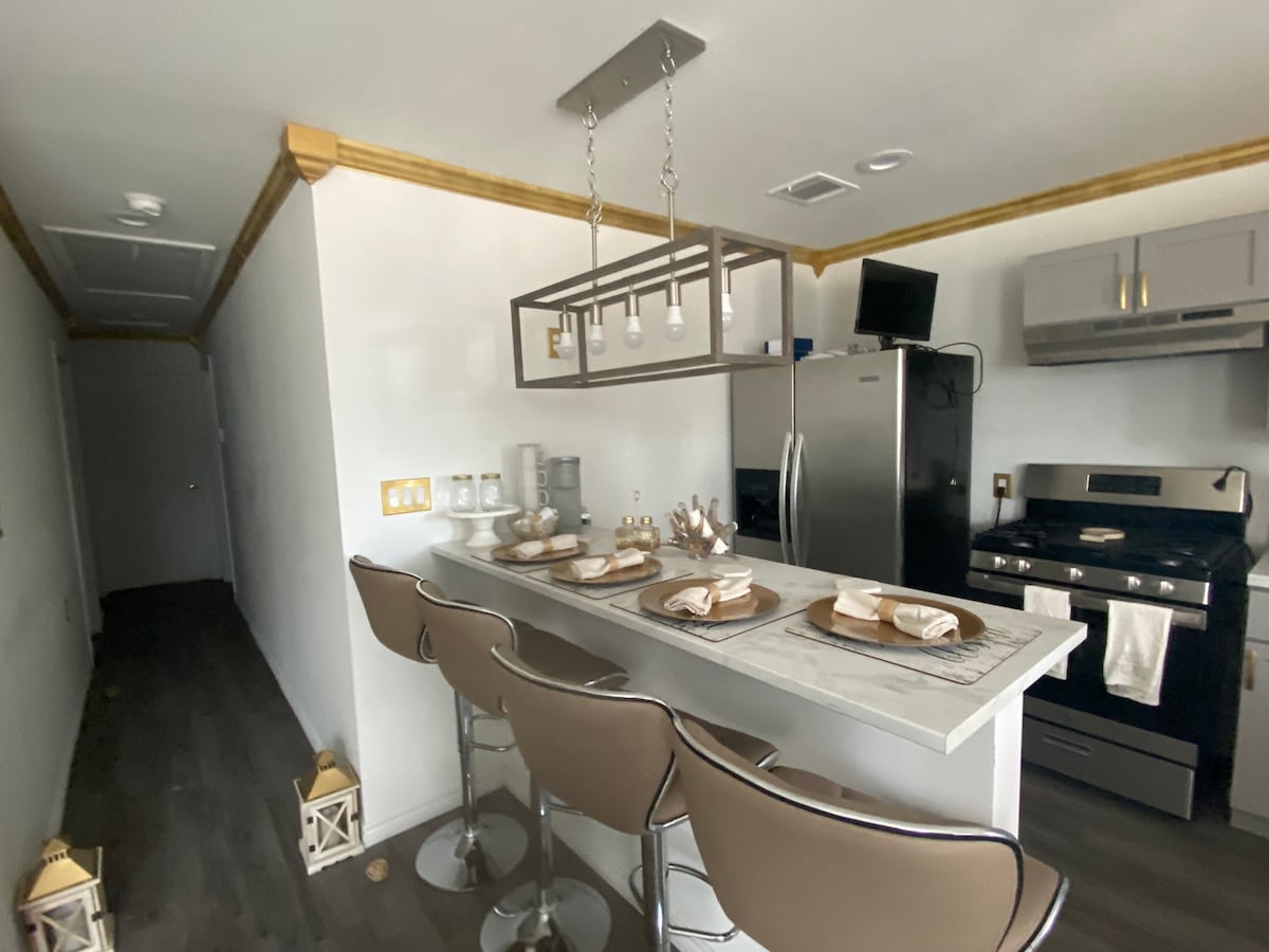 A modern kitchen features sleek cabinetry and stainless steel appliances, including a refrigerator and gas range. Three high-backed stools are positioned at a marble countertop, set with tasteful tableware. A pendant light fixture above adds a contemporary touch to the well-lit space.