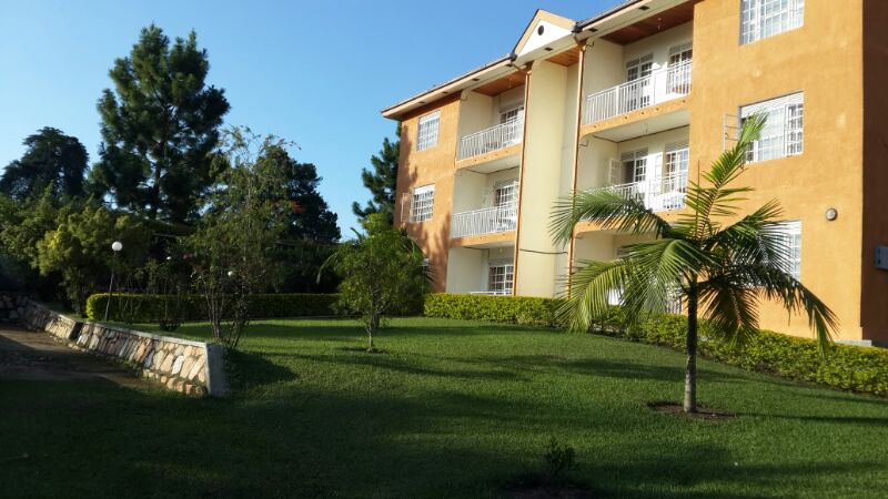 A three-story building features a warm orange exterior with multiple balconies. Lush green lawns surround the structure, accentuated by small trees and palm plants. The clear sky above provides natural light to the well-kept area, enhancing the serene atmosphere.