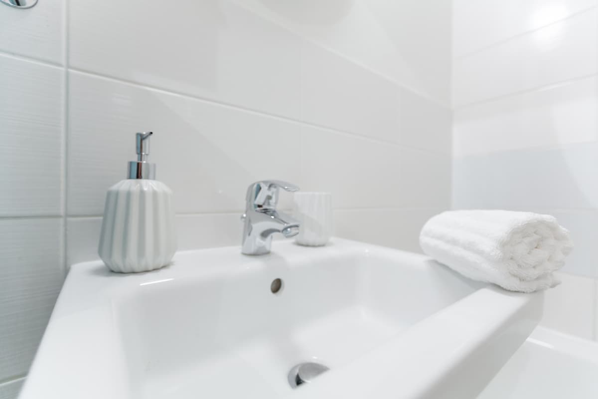 A clean bathroom sink area is presented with a sleek chrome faucet and stylish white accessories, including a soap dispenser and a toothbrush holder. A neatly rolled white towel is placed next to the sink, enhancing the serene atmosphere.