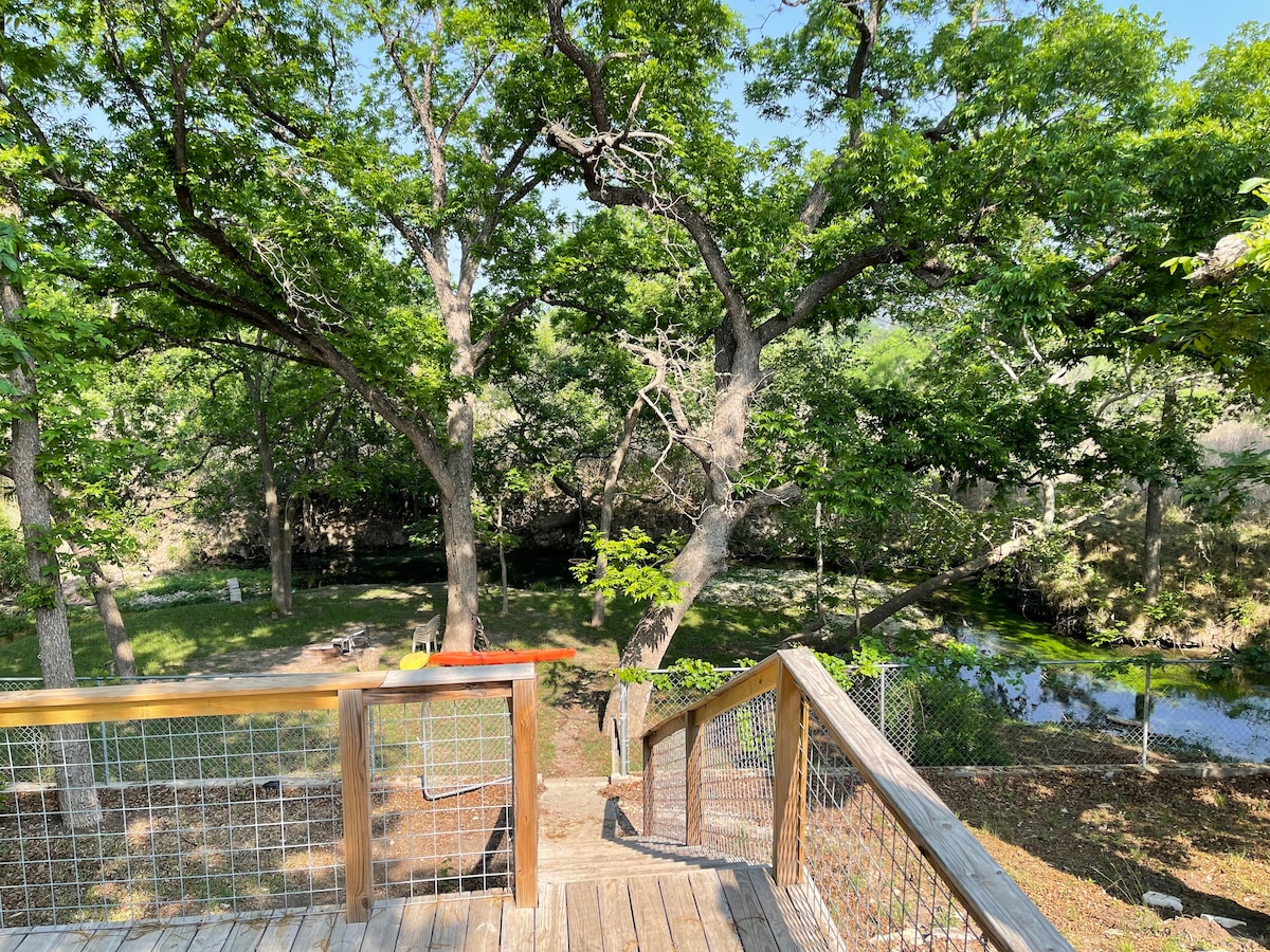 A wooden deck provides a view of lush greenery and mature trees surrounding the property. A peaceful river can be seen flowing nearby, enhancing the tranquil outdoor setting.