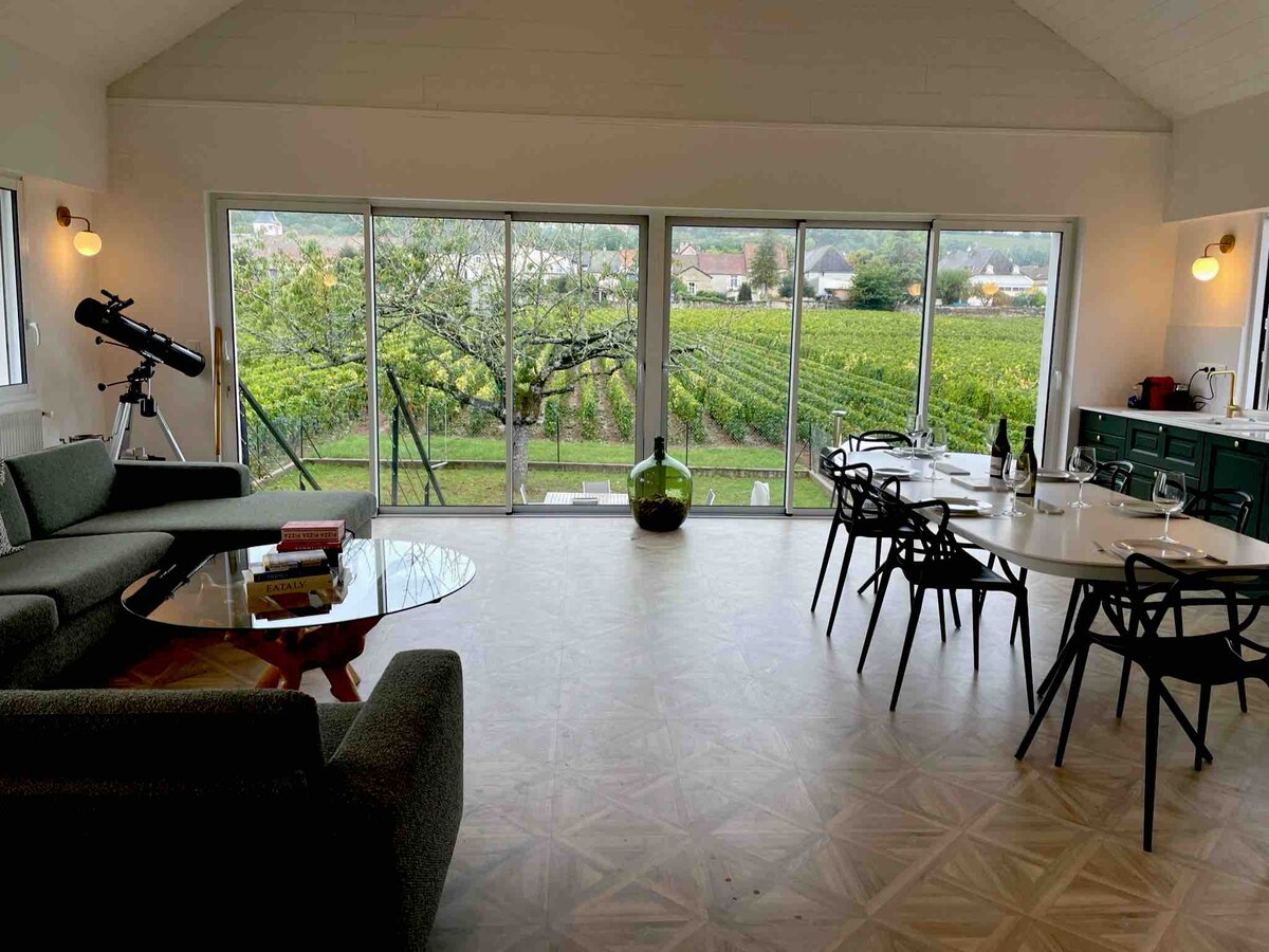 The spacious living area features large glass doors that open to vineyard views, allowing natural light to fill the room. A comfortable seating area with a dark green couch surrounds a glass coffee table. A dining table is set for several guests, with wine glasses and place settings arranged.