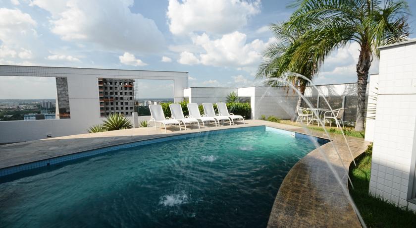 A rooftop pool area is featured, equipped with lounge chairs positioned beside the water. Lush greenery, including palm trees, adds a natural touch. The pool exhibits a clear blue hue, with water gently cascading from an integrated fountain feature.
