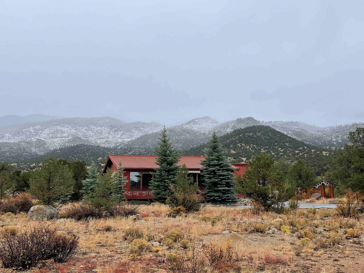 A mid-century mountain home is nestled within a natural landscape, framed by two green evergreens. The surrounding area features gentle hills and distant snow-capped peaks under a cloudy sky, with a textured foreground of dry grass and shrubs.