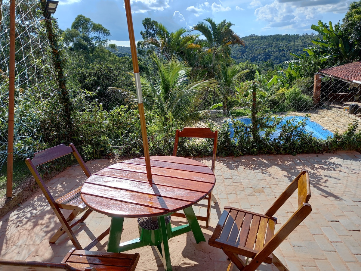 A round wooden table is surrounded by four chairs, set against a backdrop of lush greenery and distant hills. A swimming pool, partially visible, reflects the sky and is framed by tropical plants, offering a serene outdoor setting.