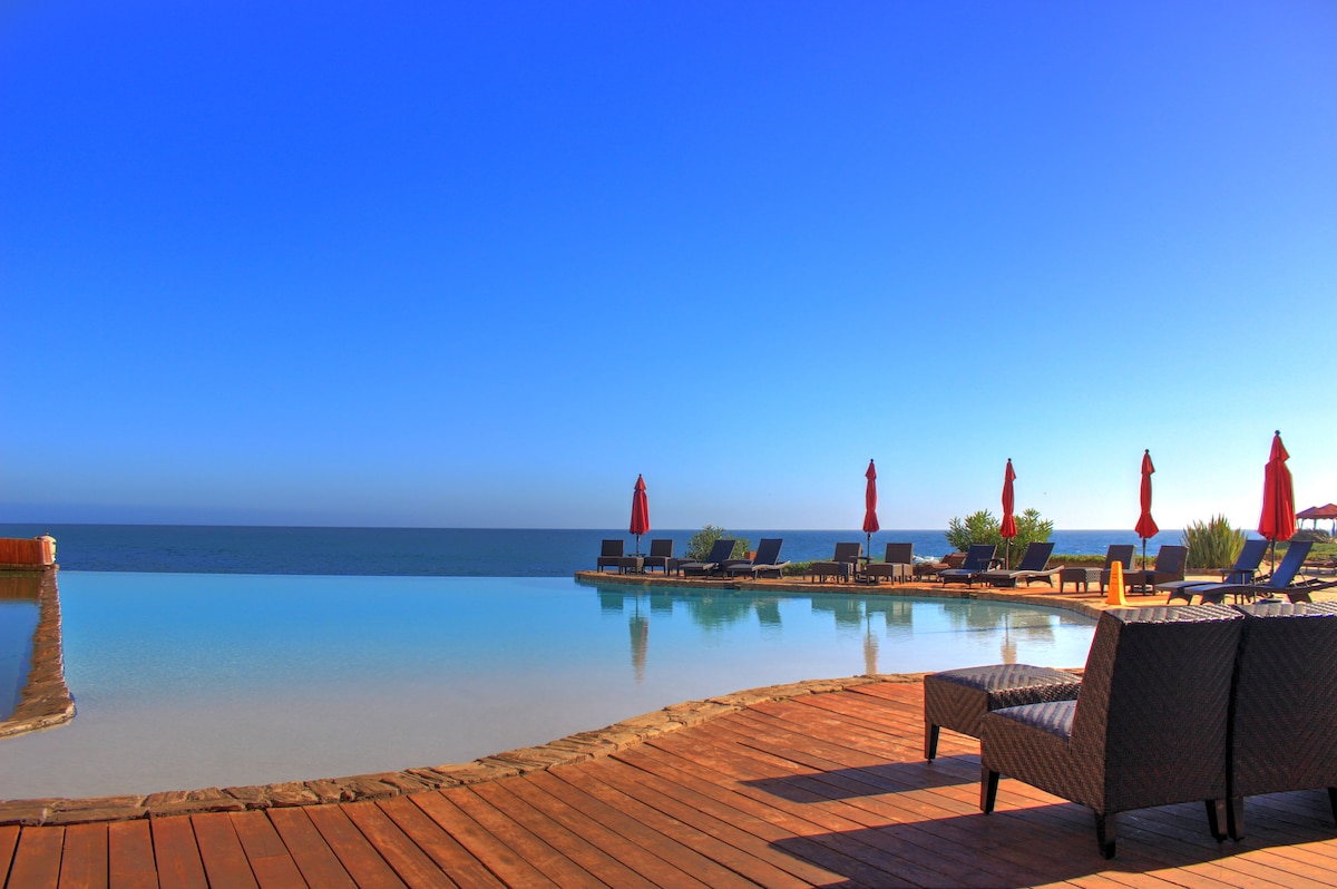 An infinity pool borders the ocean with lounge chairs placed around its edge. Red umbrellas provide shade for sunbathers. The calm water reflects the clear blue sky, creating a serene atmosphere for relaxation.