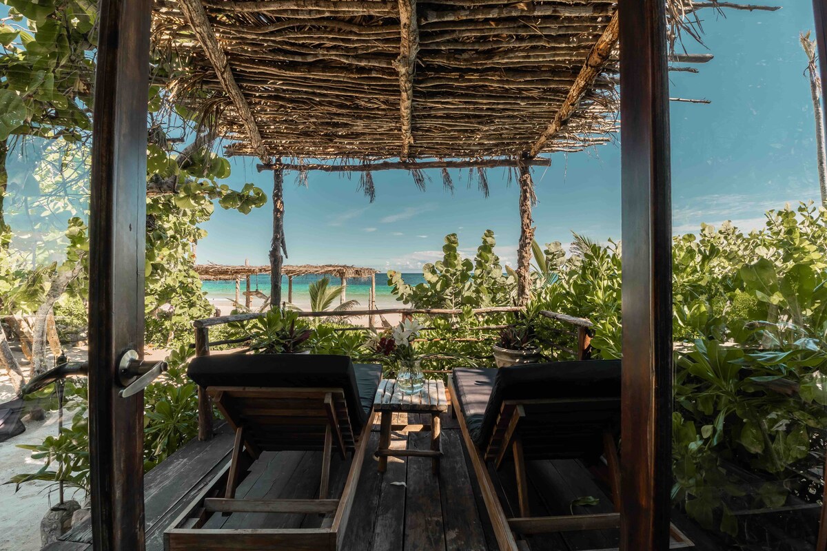 A private terrace is viewed through wooden doors, featuring lounge beds positioned for sea views. Lush greenery surrounds the space, creating a natural and peaceful ambiance, while the horizon displays calm ocean waters under a clear sky.