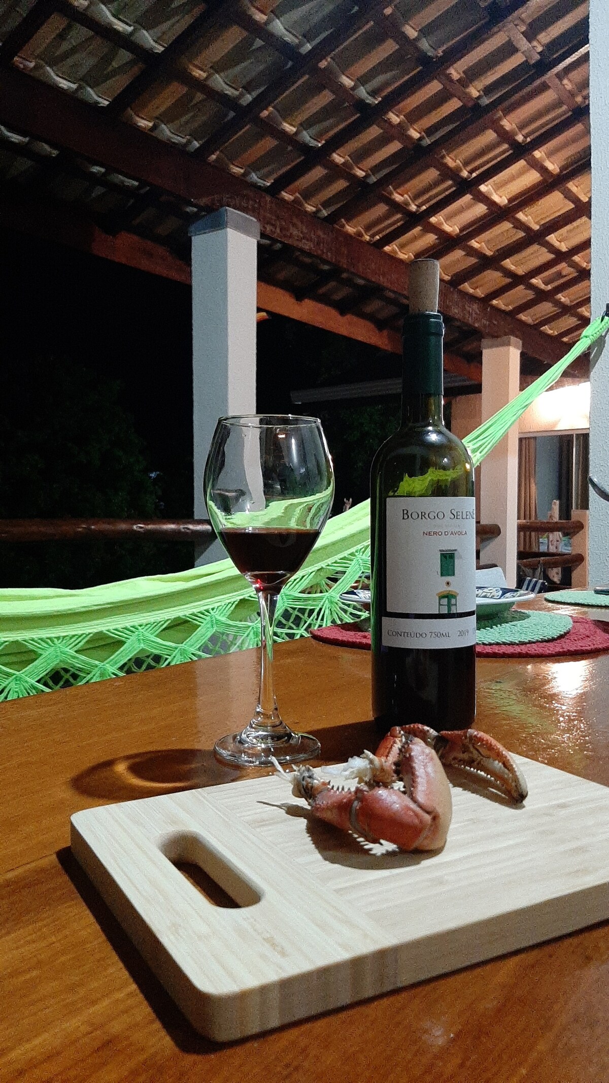 A wooden table features a glass of red wine next to a bottle on a cutting board with a shellfish. The background shows a porch area with a hammock and the soft glow of indoor lighting, creating a relaxed evening ambiance.