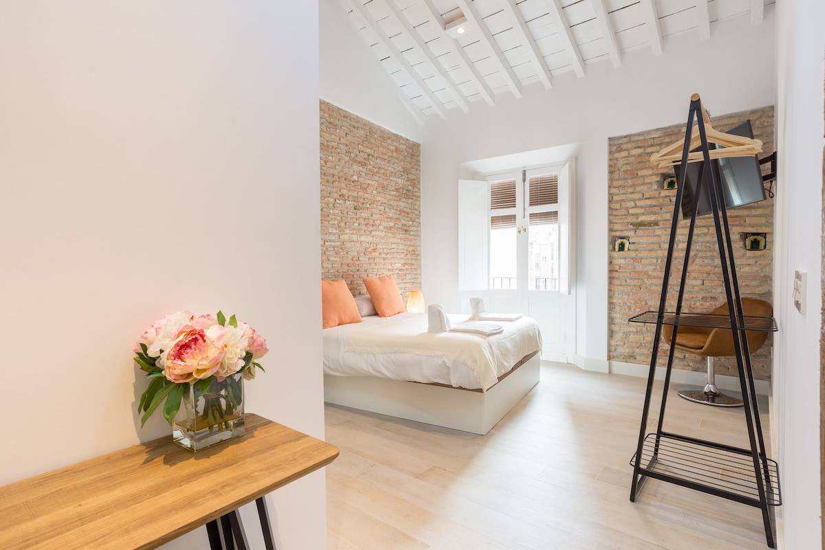 A bright bedroom features a spacious bed with soft linens, complemented by peach-colored pillows. Exposed brick walls add texture, while natural light enters through two windows. A small table with a decorative flower arrangement and a stylish clothing rack complete the inviting space.