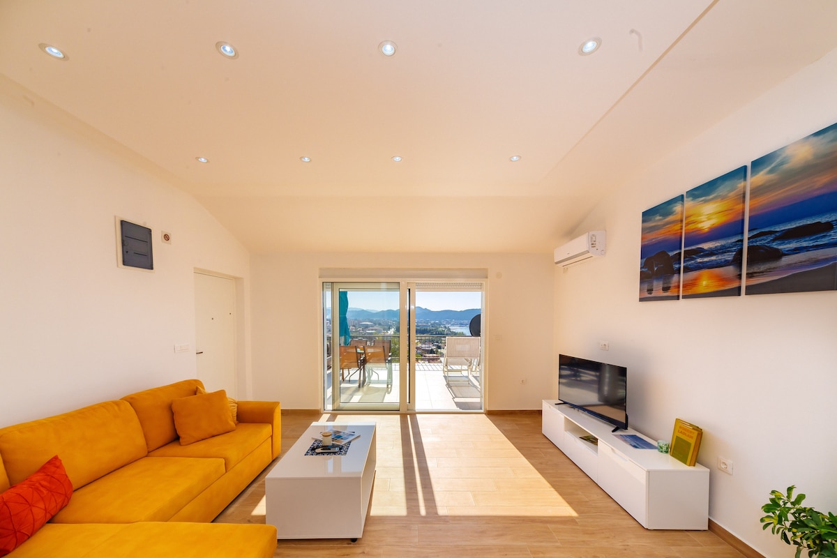 A bright living room features a yellow sectional sofa, accompanied by a sleek white coffee table. Two large glass doors provide access to a terrace, allowing natural light to fill the room. A flat-screen TV is mounted on a white entertainment unit, complemented by vibrant artwork on the walls.