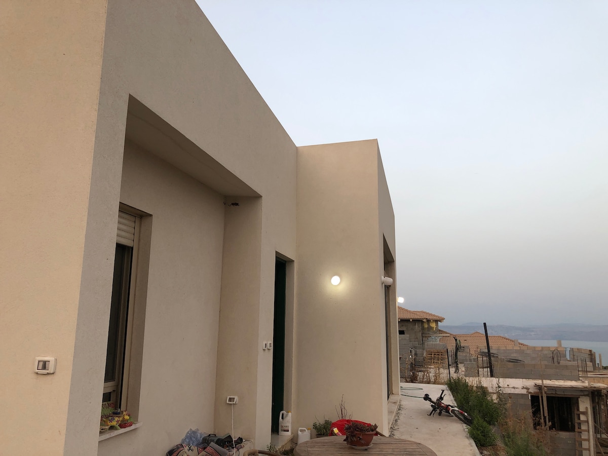 A modern building is presented, featuring clean lines and minimalistic design. Windows and exterior lights provide subtle illumination. A shaded outdoor seating area is visible, accompanied by scattered potted plants. The surrounding landscape appears open, with distant structures and an expansive view.