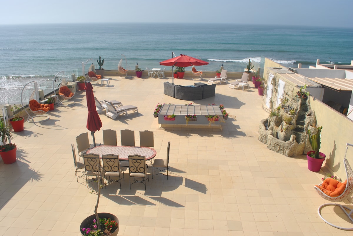 A spacious terrace offers panoramic ocean views, furnished with various seating arrangements. A large dining table accommodates up to ten guests, while lounge areas feature stylish chairs and an umbrella for shade. A jetted spa, decorative plants, and a BBQ area enhance the outdoor space.
