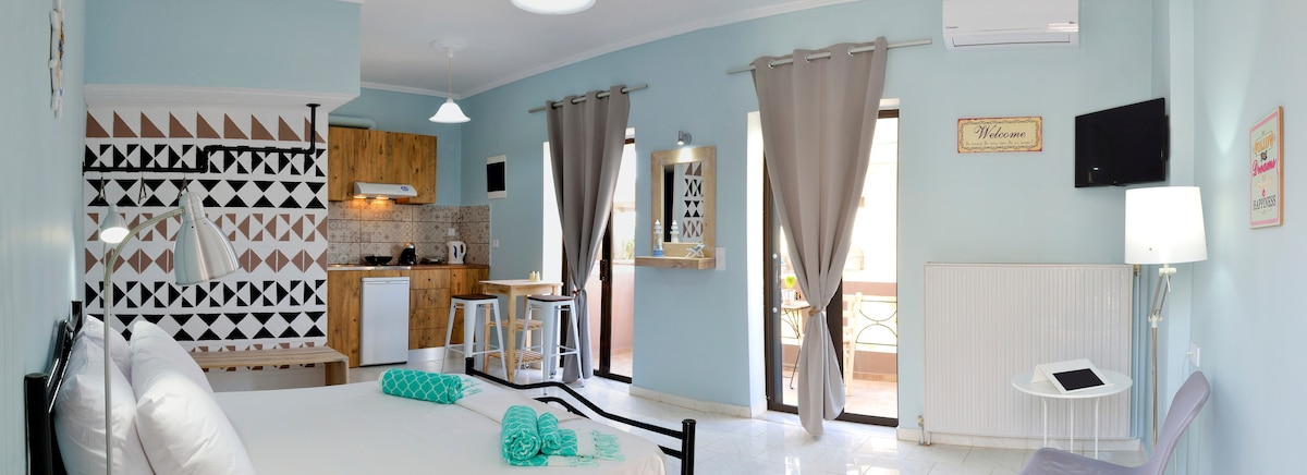 The well-appointed studio features light blue walls and a modern kitchen area with wooden cabinetry. A spacious bed is situated in the foreground, with an inviting view of the balcony seen through large glass doors. Natural light fills the room, complemented by stylish decor.