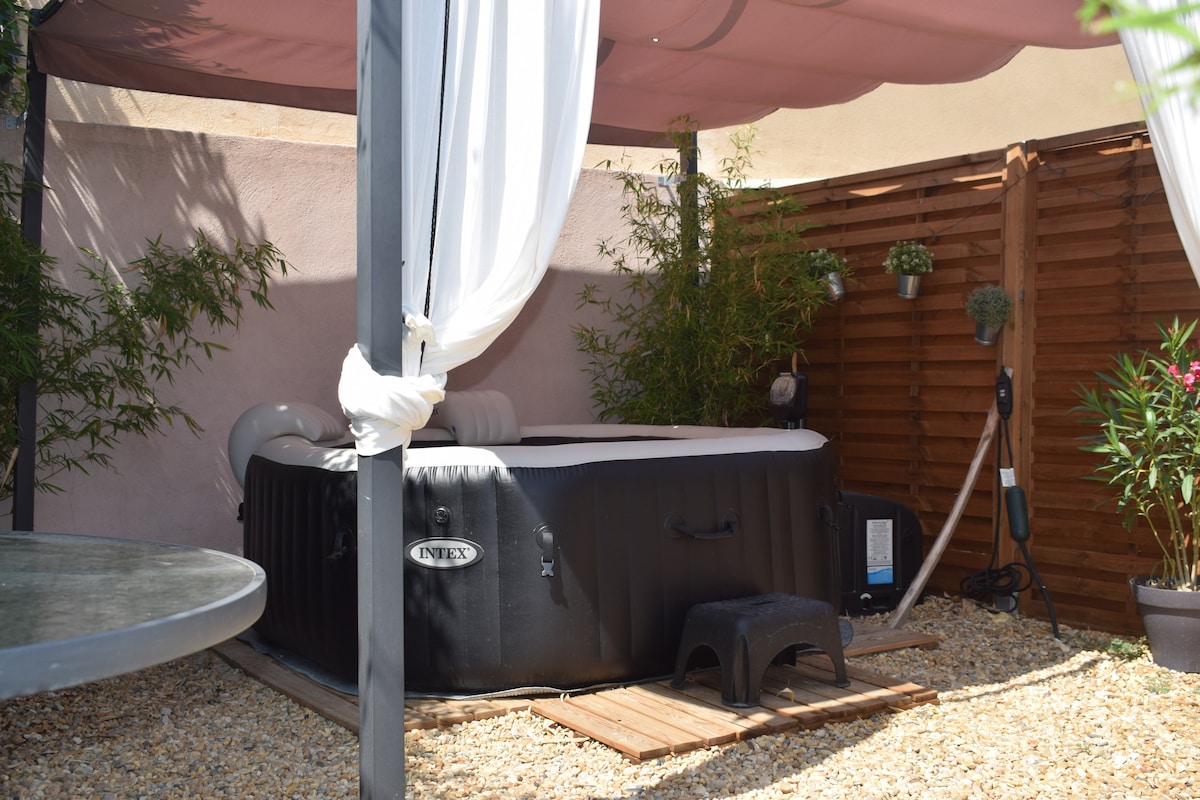 An outdoor spa area is displayed, featuring a portable hot tub surrounded by privacy curtains and wooden fencing. Potted plants and bamboo contribute to a serene ambiance, while a small wooden platform and a step stool provide easy access. A table is partially visible in the foreground.
