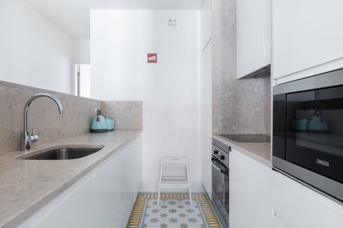 A modern kitchen features sleek white cabinetry and a light stone countertop. A sink is positioned on the left, with a kettle and small appliances on display. The colorful patterned tile floor adds a touch of charm, and a small step stool is visible near the oven.