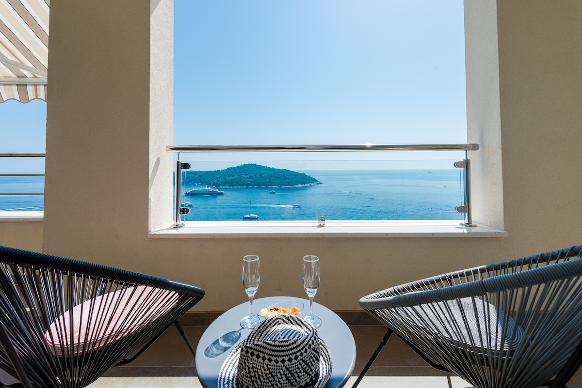 A private balcony offers a stunning view of the sea and the island of Lokrum. Two stylish chairs face a round table, which holds small snacks and two glasses, inviting relaxation while enjoying the scenery. Bright sunlight enhances the serene atmosphere.