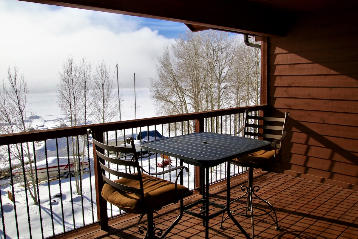 A private balcony features a small dark table and two chairs, each with cushions, overlooking a snowy landscape. Bare trees and distant boats are visible, framed by the wooden structure of the balcony. Soft clouds cloak parts of the view.
