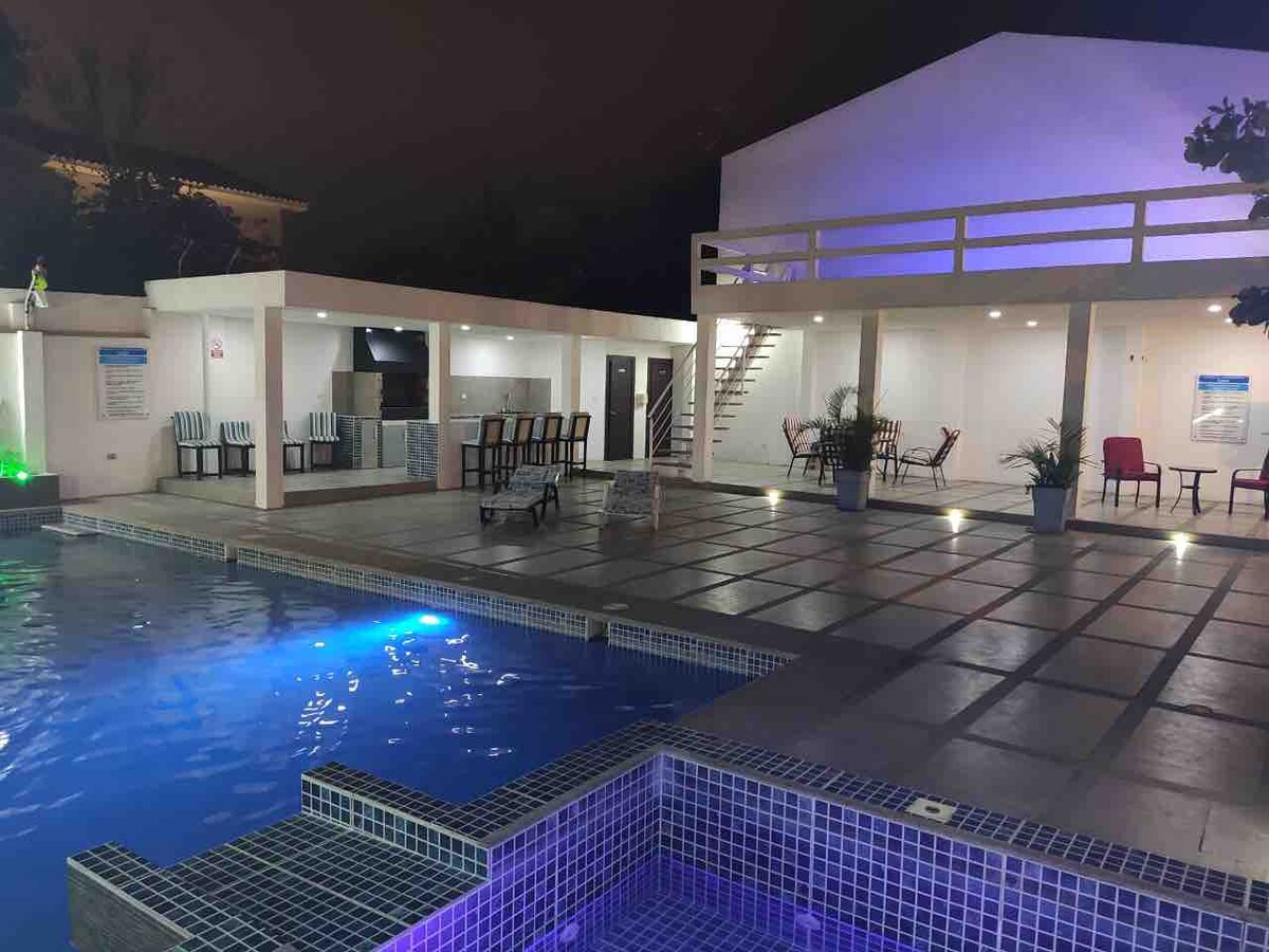 A communal area is highlighted by a large swimming pool, featuring illuminated blue tones. Surrounding spaces include multiple seating options, dining tables, and a jacuzzi. The two-story building is visible in the background, with stairs leading to an upper level.
