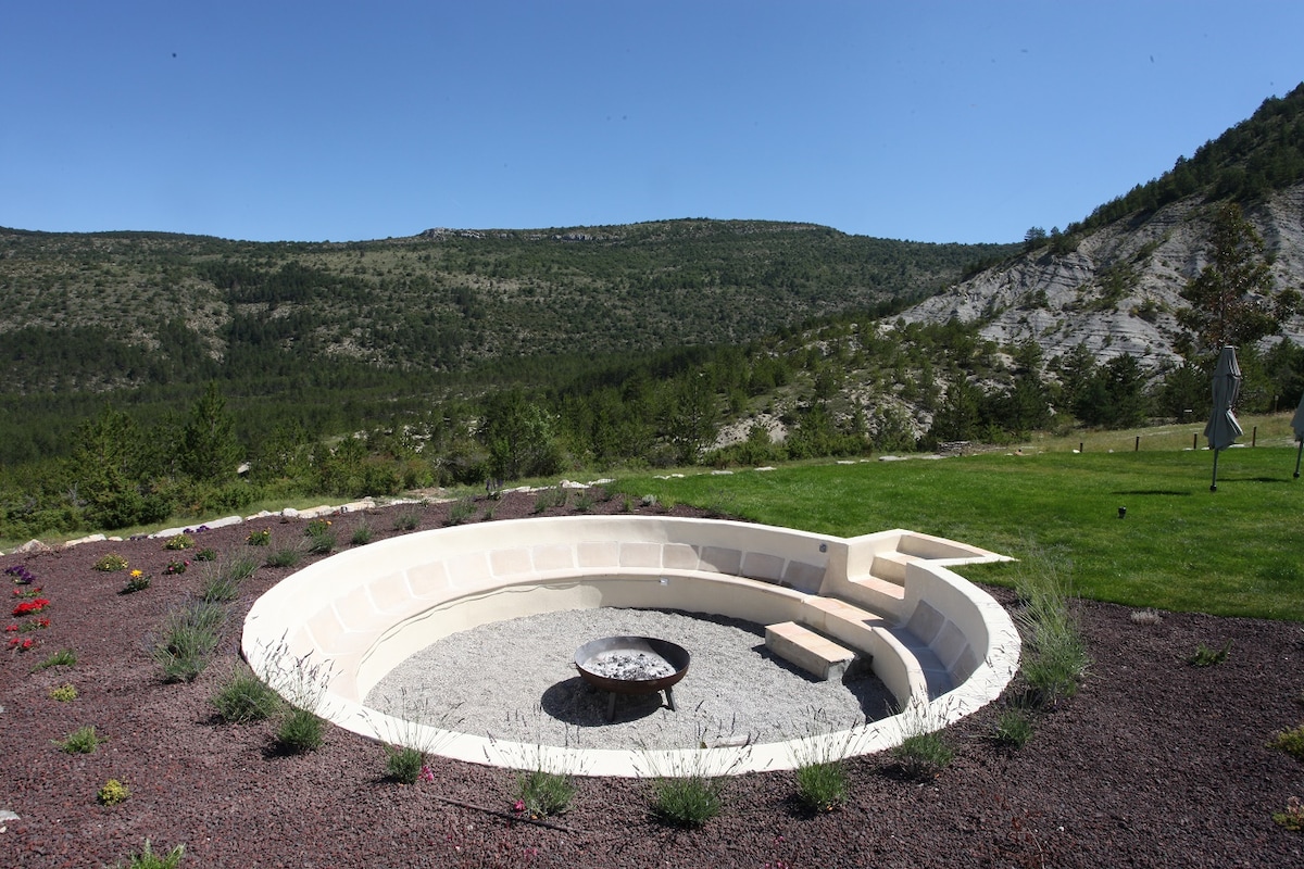 A circular fire pit area is surrounded by a landscaped garden featuring low plants and pebbles. The scenic backdrop includes green hills and rocky cliffs under a clear blue sky, creating a serene outdoor setting for gatherings.
