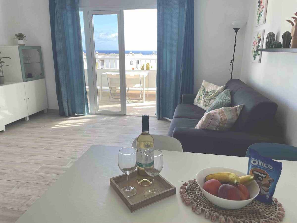 A bright living area features a comfortable dark gray sofa and a dining table set with glasses and a bottle of wine. A bowl of fruit rests on the table. The large glass doors open to a balcony with a view of the ocean.