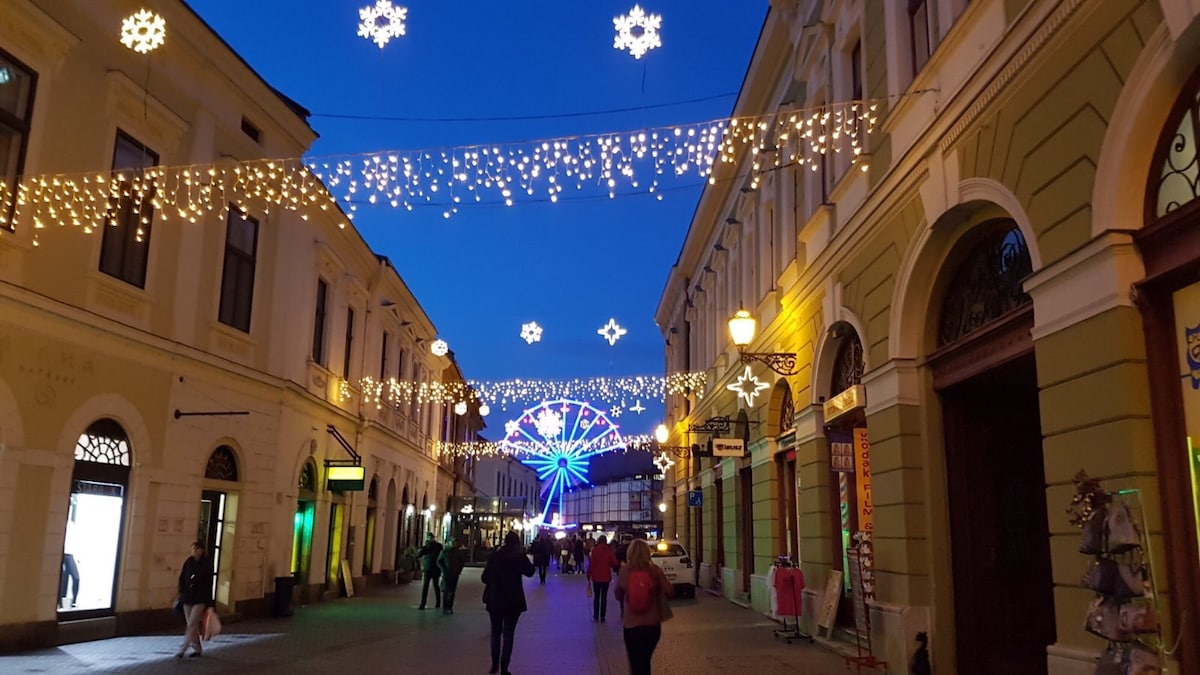 A lively pedestrian street is showcased, adorned with twinkling lights and decorative snowflakes. Colorful illumination from a large display at the end of the street creates a festive atmosphere, while people stroll leisurely, enjoying the vibrant ambiance of the surrounding historic architecture.