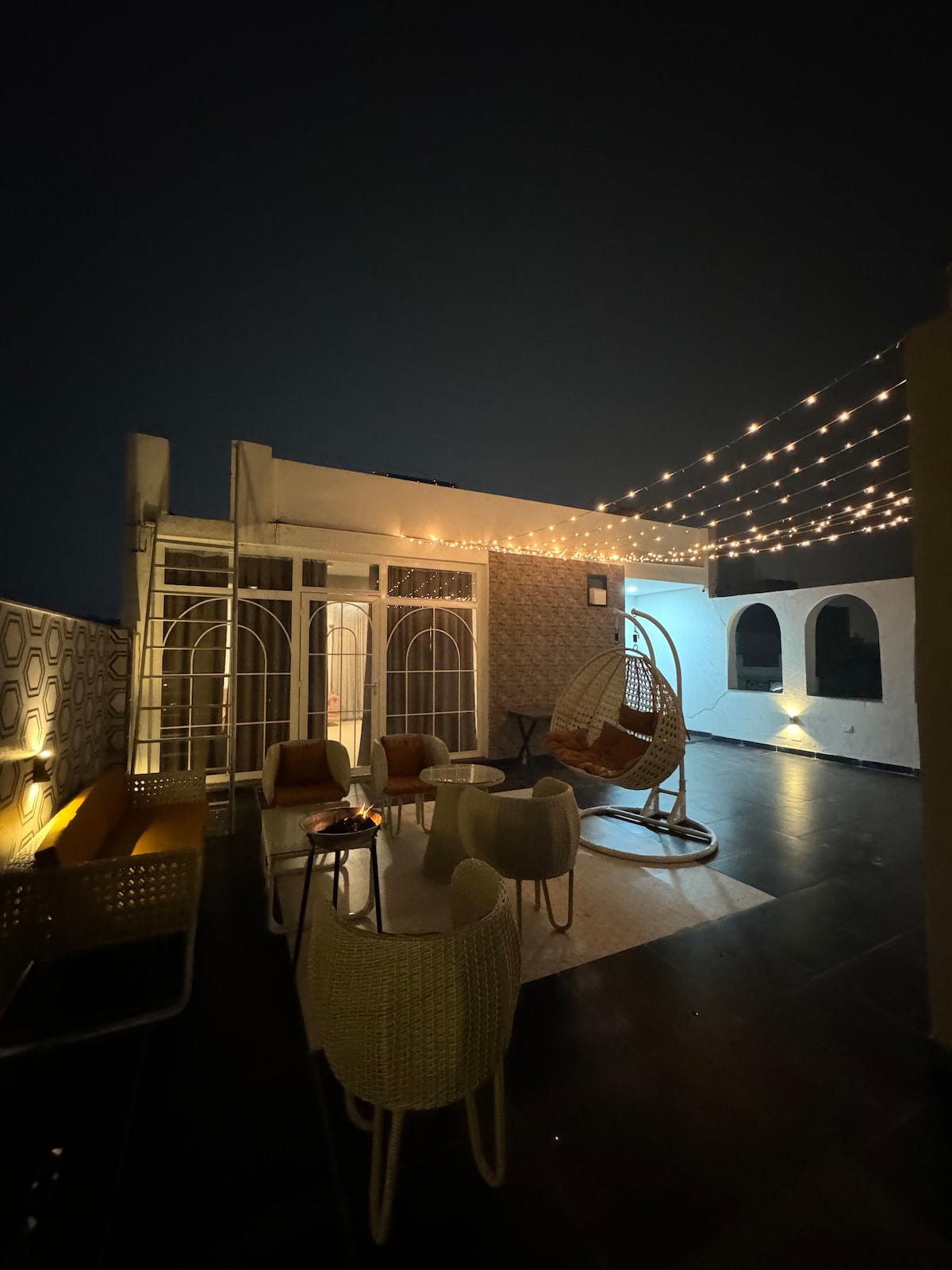 A rooftop area is illuminated by warm string lights, creating a cozy ambiance. Comfortable seating options include hanging chairs and soft lounge seating arranged around a central table. The backdrop features modern architectural elements, enhancing the inviting space.