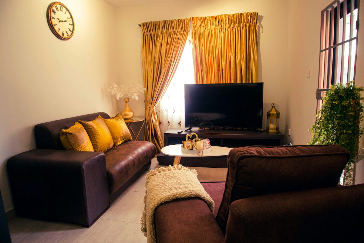 A cozy living area features two sofas adorned with golden cushions, complemented by a coffee table. A large television is positioned against the wall, while elegant golden curtains frame a window, allowing natural light to brighten the space. A decorative plant adds a touch of greenery.