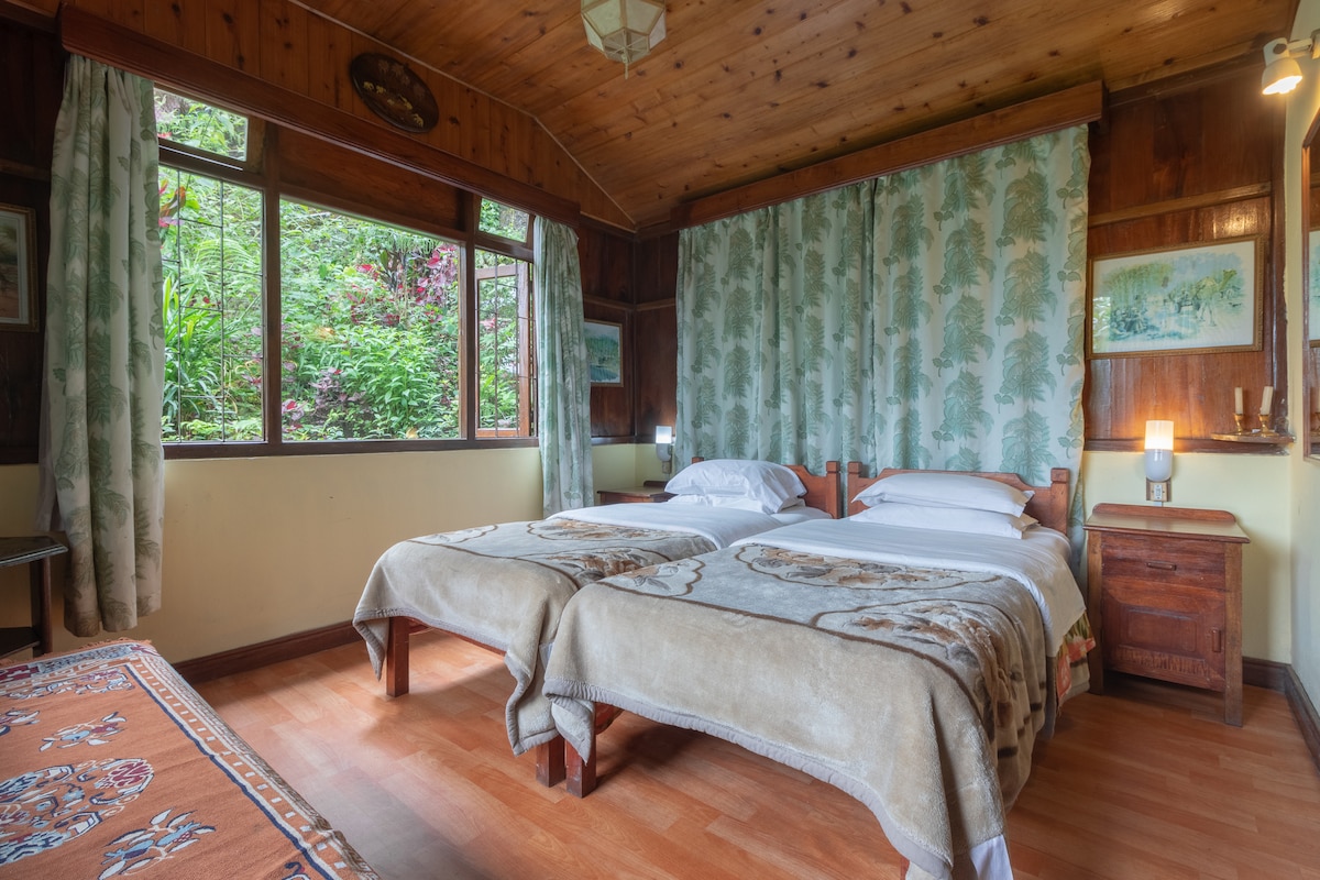 A cozy bedroom is presented, featuring two spacious beds adorned with soft blankets. Natural light filters through large windows, offering views of the surrounding greenery. Wooden accents and nature-inspired curtains enhance the room's serene ambiance.