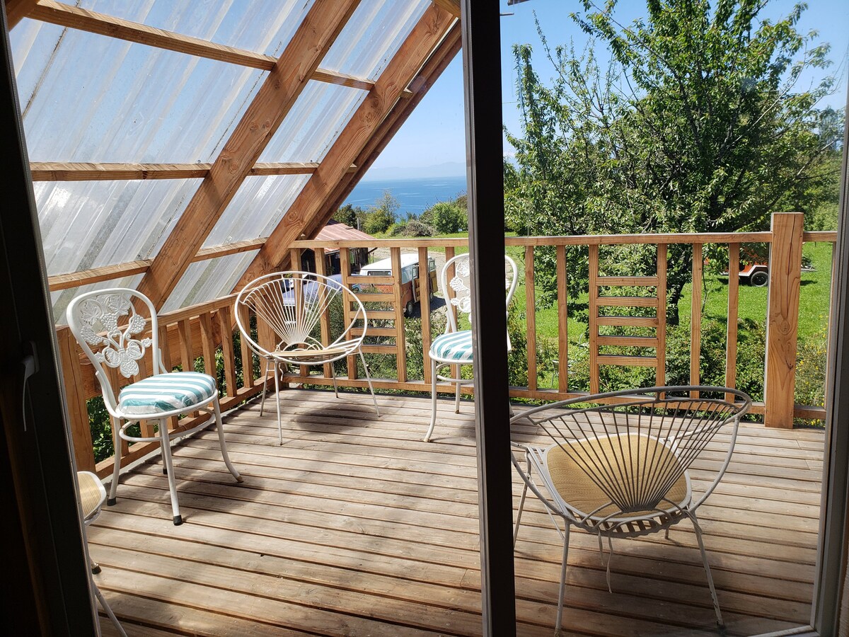 A wooden deck is featured with multiple seating options, including two stylish chairs with circular designs. The enclosure showcases transparent roofing, allowing natural light to flood the area. Lush greenery and distant views of the ocean are visible beyond the railing.