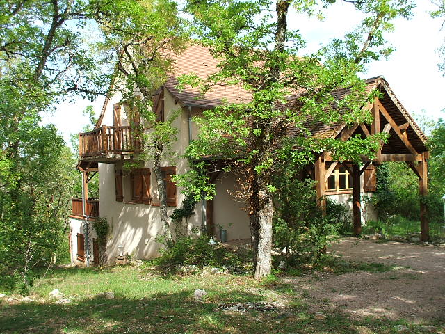 A charming vacation house is surrounded by lush greenery and trees. The two-story structure features wooden shutters and a balcony, evoking a warm, rustic feel. A covered entrance adds to the inviting atmosphere, while the natural setting provides a sense of privacy and tranquility.