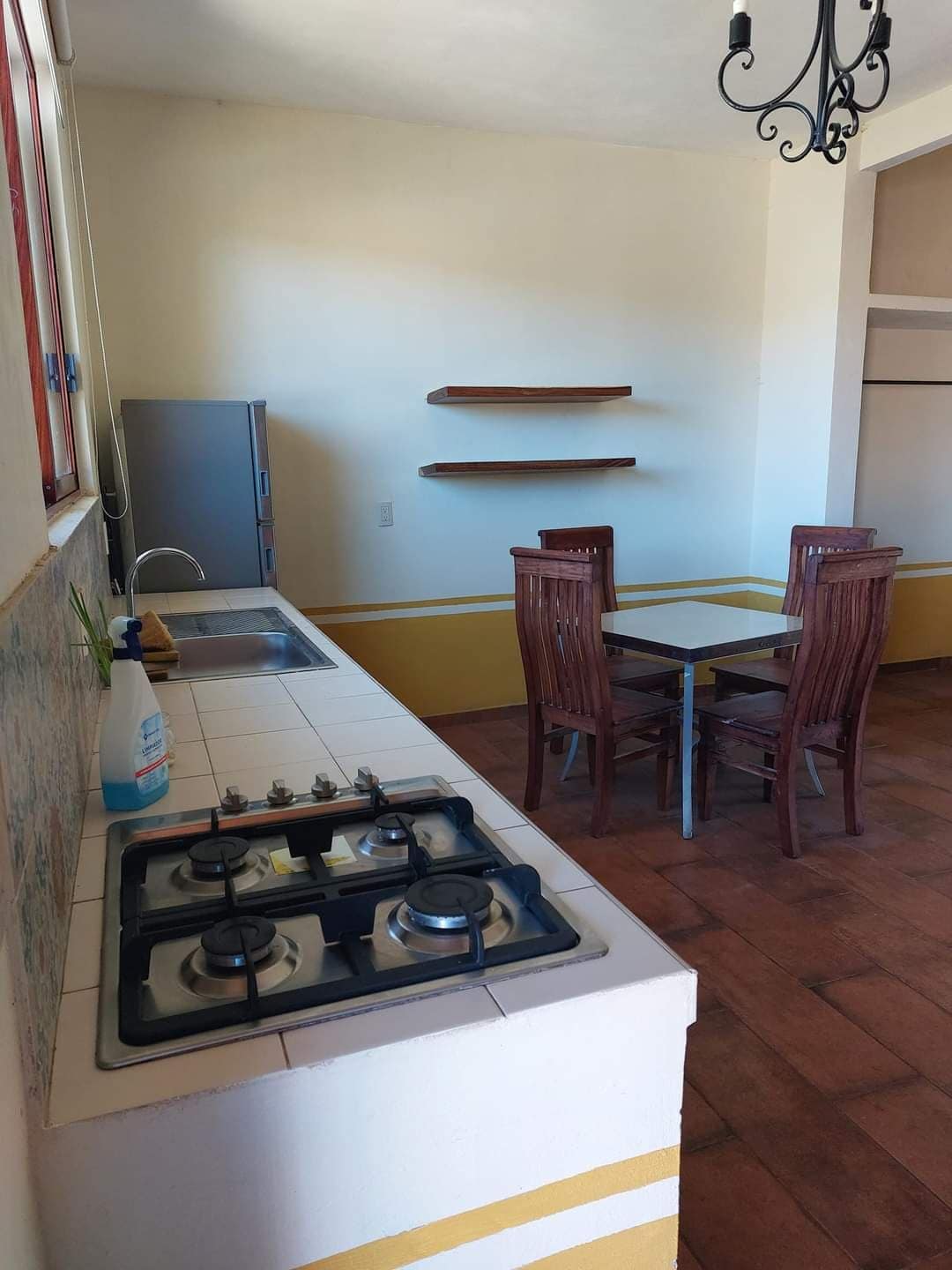 A functional kitchen area features a gas stove with multiple burners and a stainless steel refrigerator. A small dining table is surrounded by four wooden chairs, and open shelving is mounted on the wall, offering space for storage or display.
