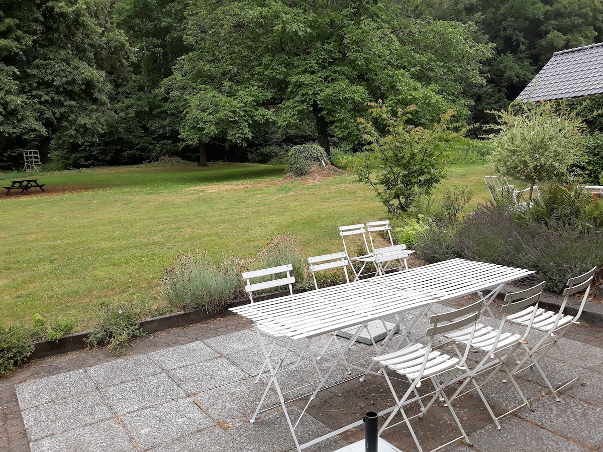 An outdoor dining area is presented with a long white table surrounded by several chairs, positioned on a stone patio. Lush greenery fills the background, featuring large trees and an expansive lawn. A picnic table is visible further back, adding to the serene forest setting.