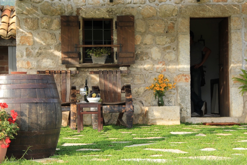 A rustic stone cottage features a welcoming entrance with wooden shutters and a potted flower arrangement. Two wooden chairs and a small table are set in front, alongside a decorative barrel. Lush greenery surrounds the outdoor space, providing a peaceful view.