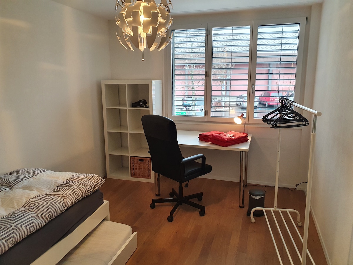 A room features a spacious bed with patterned bedding and a light wooden floor. A desk with a lamp is situated beside a window with modern blinds, while a tall shelf and a clothing rack provide additional storage. A chair completes the workspace.