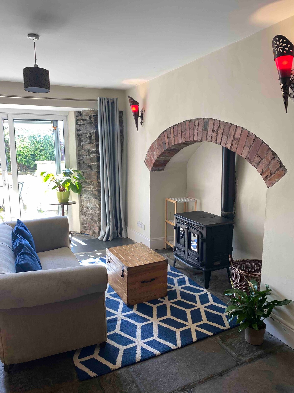 A cozy living area features a light-colored sofa with blue cushions, set beside a wood burning stove. A coffee table is positioned in front, and natural light floods the space through the large glass doors leading to a garden. Textured stone walls add character.