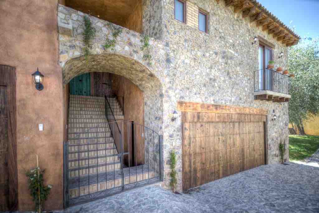 The entrance features a charming archway leading to a stone staircase, surrounded by earthy tones and natural textures. A wooden garage door complements the rustic aesthetic, while greenery can be seen along the archway, adding a touch of life to the stone facade.