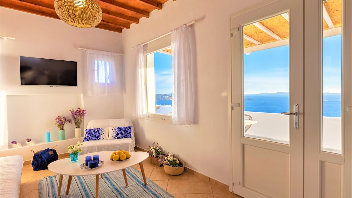 A bright living area is highlighted by natural light streaming through large windows that offer views of the Aegean Sea. A white sofa is positioned beside a decorative coffee table set with fruit. Wall-mounted TV and tasteful accents add to the inviting space.