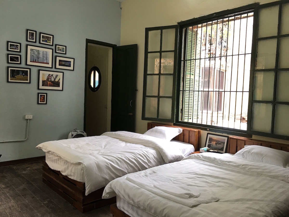 A room features two spacious beds with white linens, positioned parallel to a large window framed by green shutters. A wooden wall provides a rustic touch, and framed photographs in various sizes decorate the wall. A round door is visible in the background.