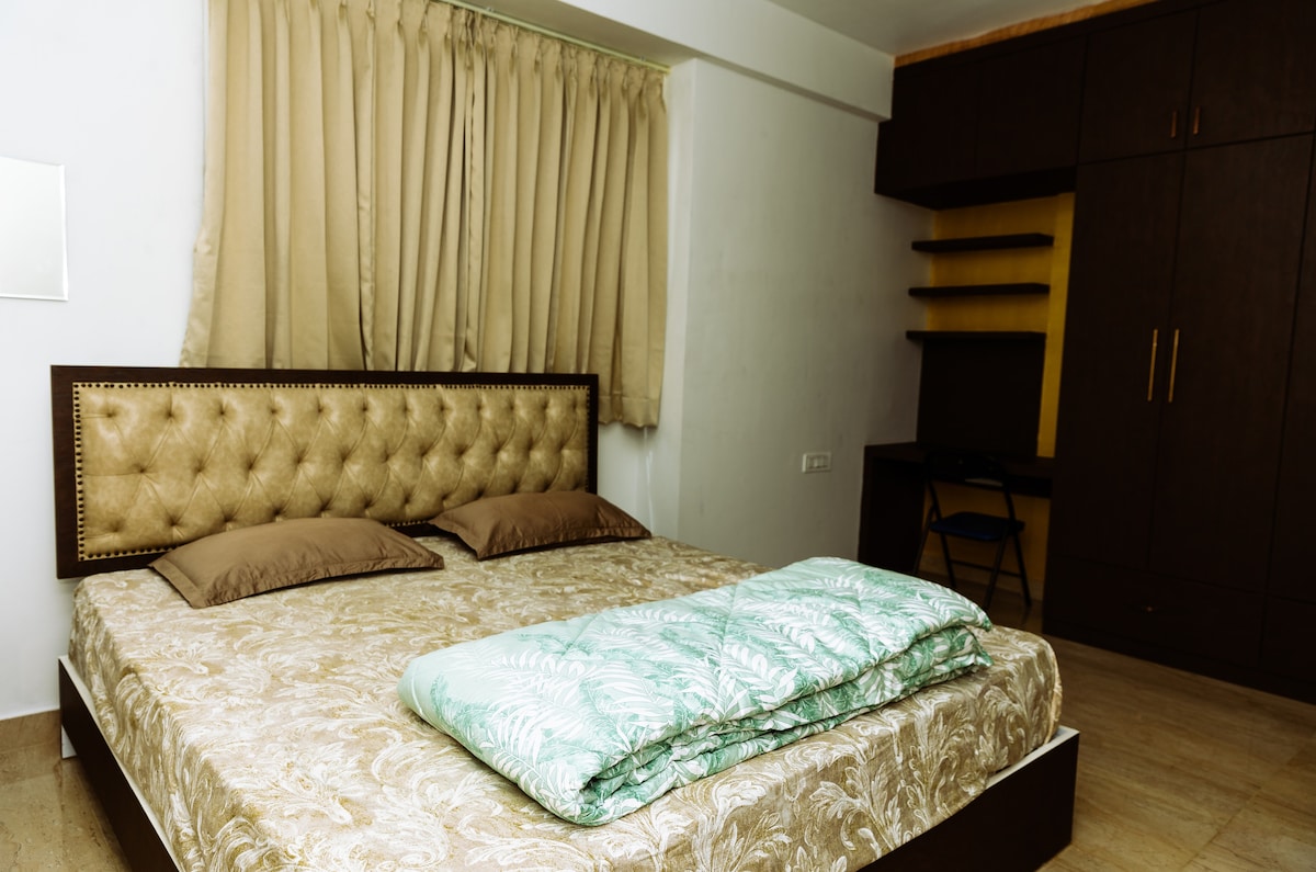 A spacious bedroom is showcased with a large bed featuring a tufted headboard. Soft brown pillows are placed on the bed, along with a patterned blanket. Natural light enters through the beige curtains, and a dark wooden wardrobe with open shelving is visible against the wall.
