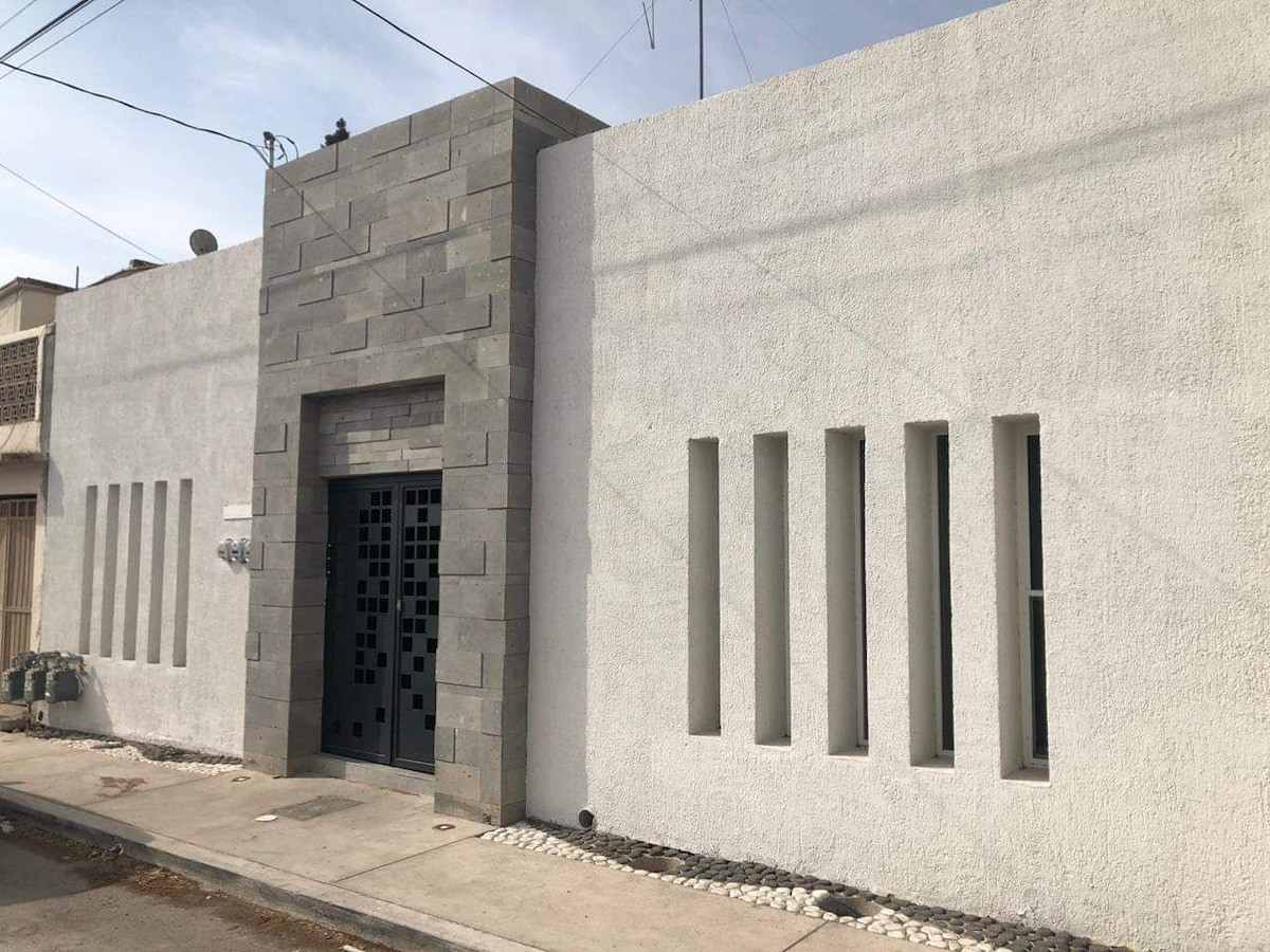 The modern exterior of the building showcases a combination of textured gray stone and smooth white walls. The entrance features an ornate black door framed by a neutral-toned facade. Vertical windows are aligned symmetrically on either side, allowing for natural light to enter the interior.