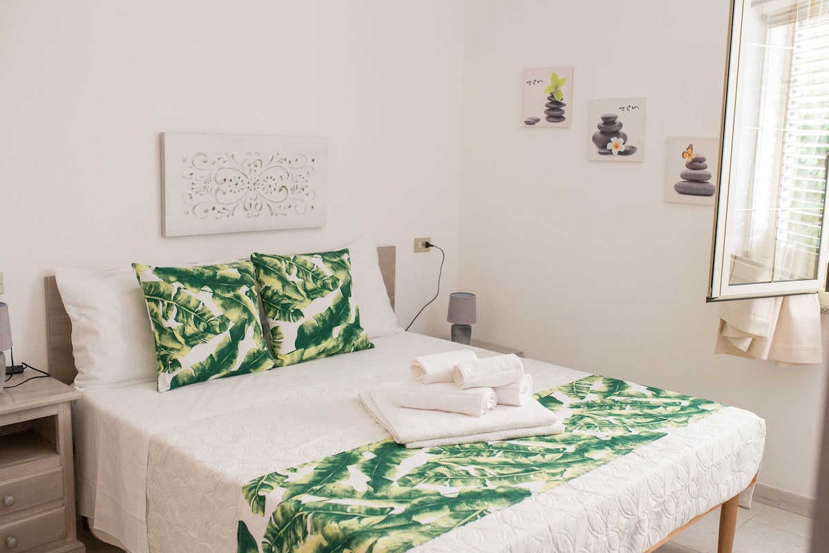 A comfortable bedroom features a spacious bed adorned with tropical patterned linens. Neatly arranged towels are placed on the bed. The room is accented by light-colored walls and decorative artwork, while a window allows natural light to enhance the serene atmosphere.