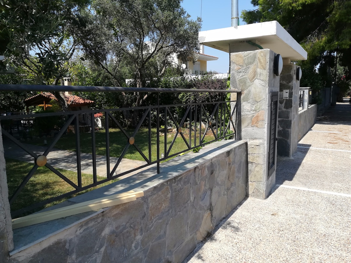 A stone wall with a decorative metal railing is featured, providing a border to the property. Lush greenery and a variety of plants are visible in the background, contributing to a serene outdoor environment. The entrance to the property is marked by a stone pillar.