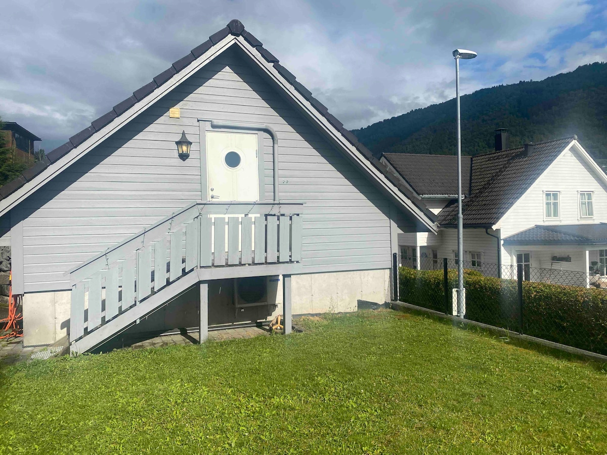 A light gray house with a sloped roof is featured, set against a backdrop of green hills. An entrance with a white door and a small staircase leads to a welcoming front porch. Nearby, another house with a dark roof is visible.
