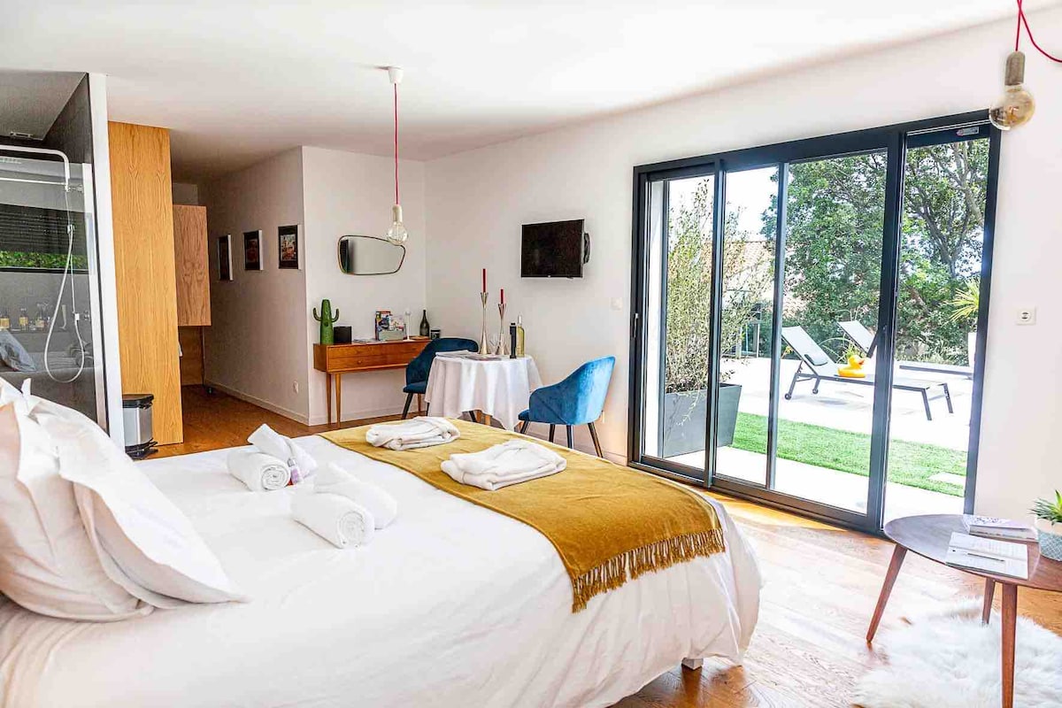 A spacious bedroom features a large bed with neatly arranged towels. A sliding glass door allows natural light to fill the room, leading to an outdoor area with lounge chairs and greenery. Soft furnishings in blue complement the warm wooden tones of the floor.