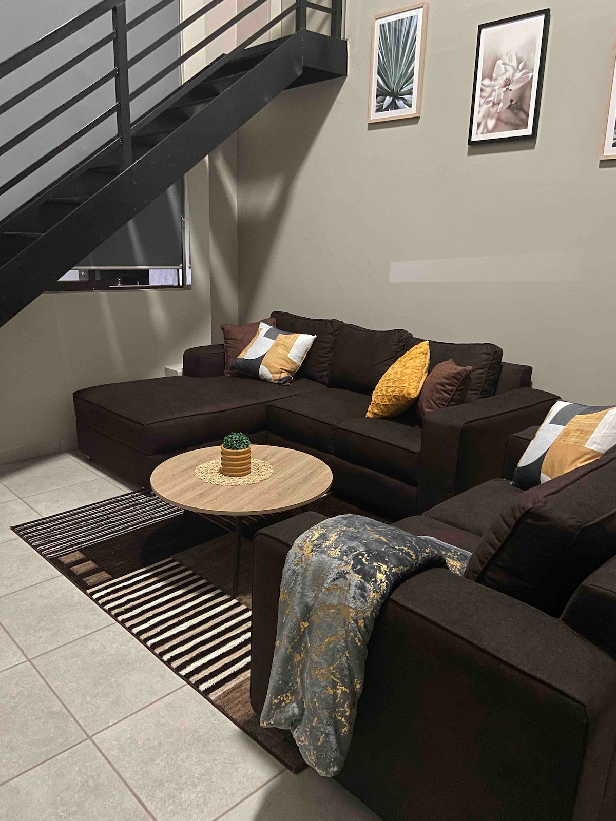 A cozy living area is presented with a dark sectional sofa adorned with decorative cushions in warm tones. A round coffee table sits in the center, complemented by a textured area rug and a soft throw draped over the armrest.