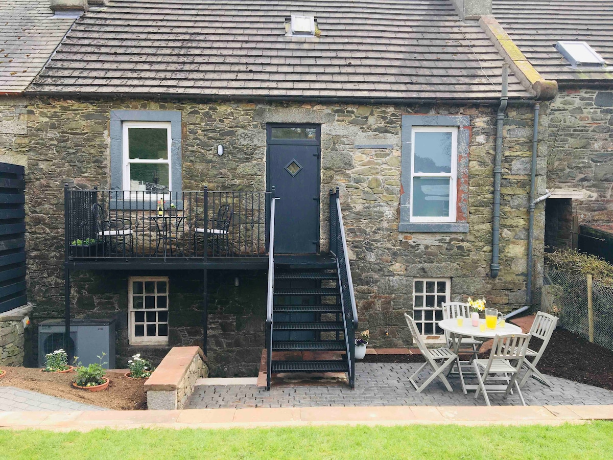 The exterior of the stone cottage features a set of stairs leading to a balcony. A patio area with a round table and multiple chairs is positioned in front, surrounded by a lush green lawn and garden beds.