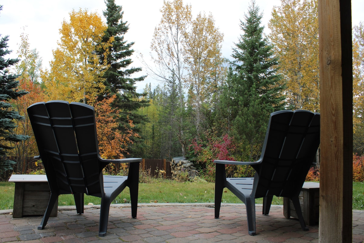 Two outdoor chairs are positioned on a stone patio, offering a view of the encircling trees in autumn foliage. The surrounding landscape features a variety of trees, showcasing vibrant colors during the fall season.